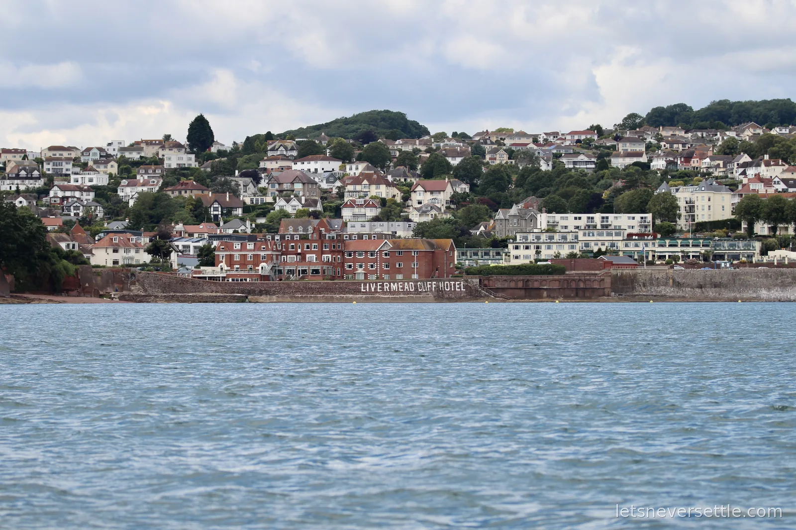 The red-brick Livermead Cliff Hotel on the Torquay seafront with its name painted along the sea wall and houses climbing the wooded hill behind