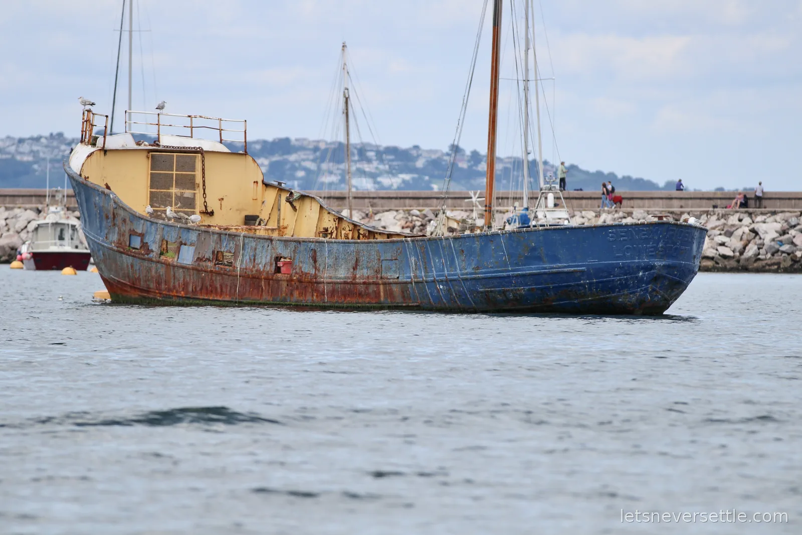 A rusting blue and yellow trawler at anchor with seagulls perched on the rigging and a stone breakwater behind