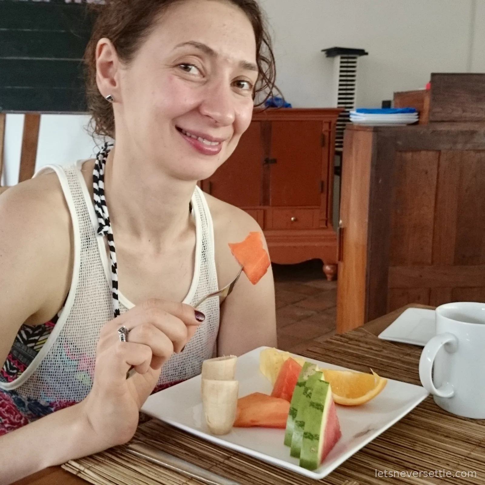 Inga at a wooden table lifting a forkful of papaya from a plate of banana, watermelon, papaya and orange