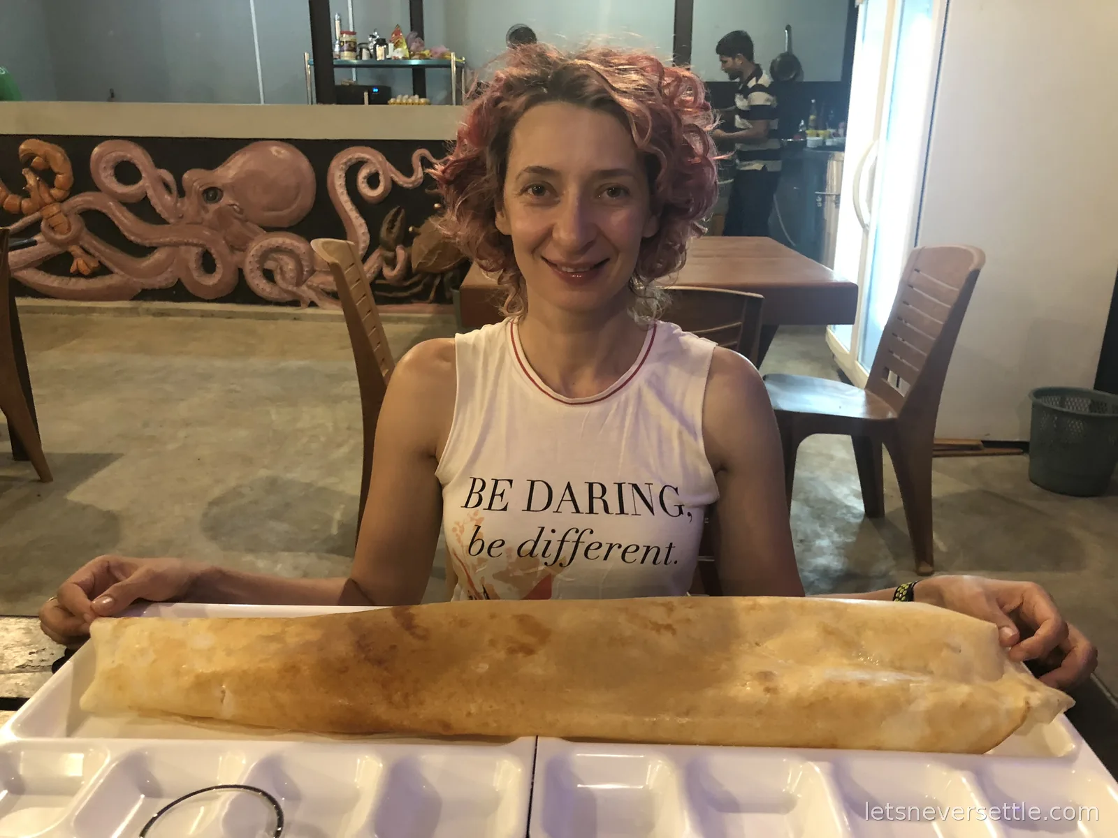 Inga smiling behind a paper dosa nearly a metre long served on two trays pushed together, an octopus mural on the wall behind