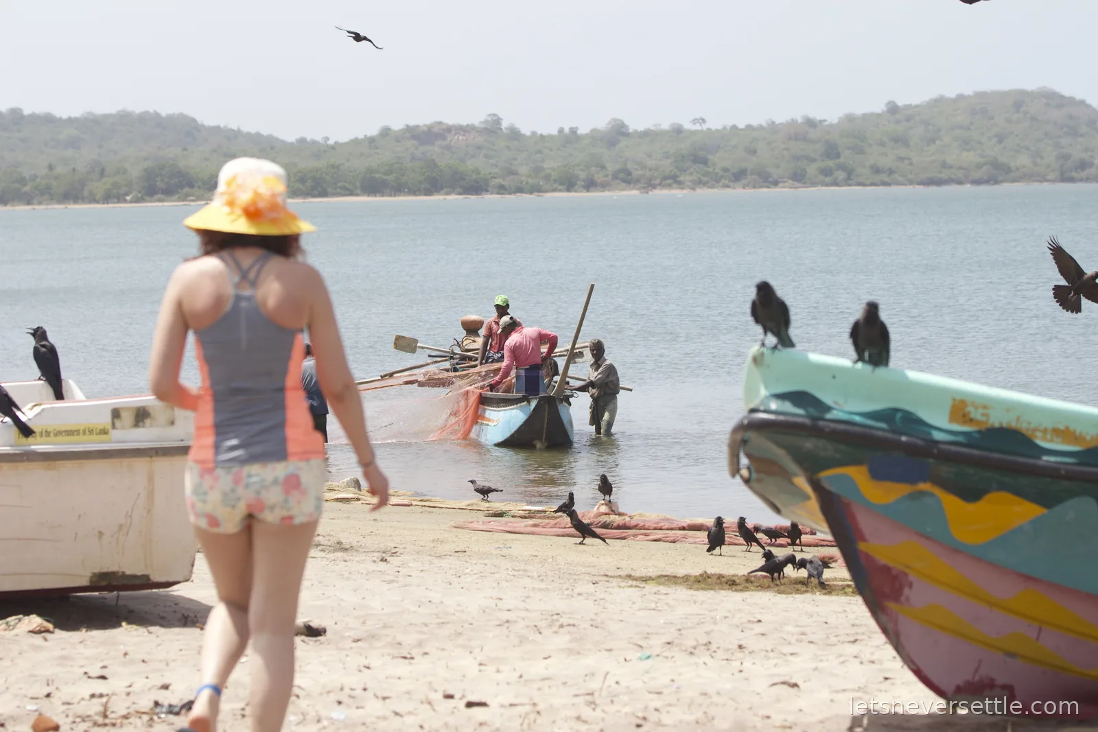 Inga in a yellow sun hat walking towards fishing boats at the water's edge, dozens of black crows perched on the painted hulls