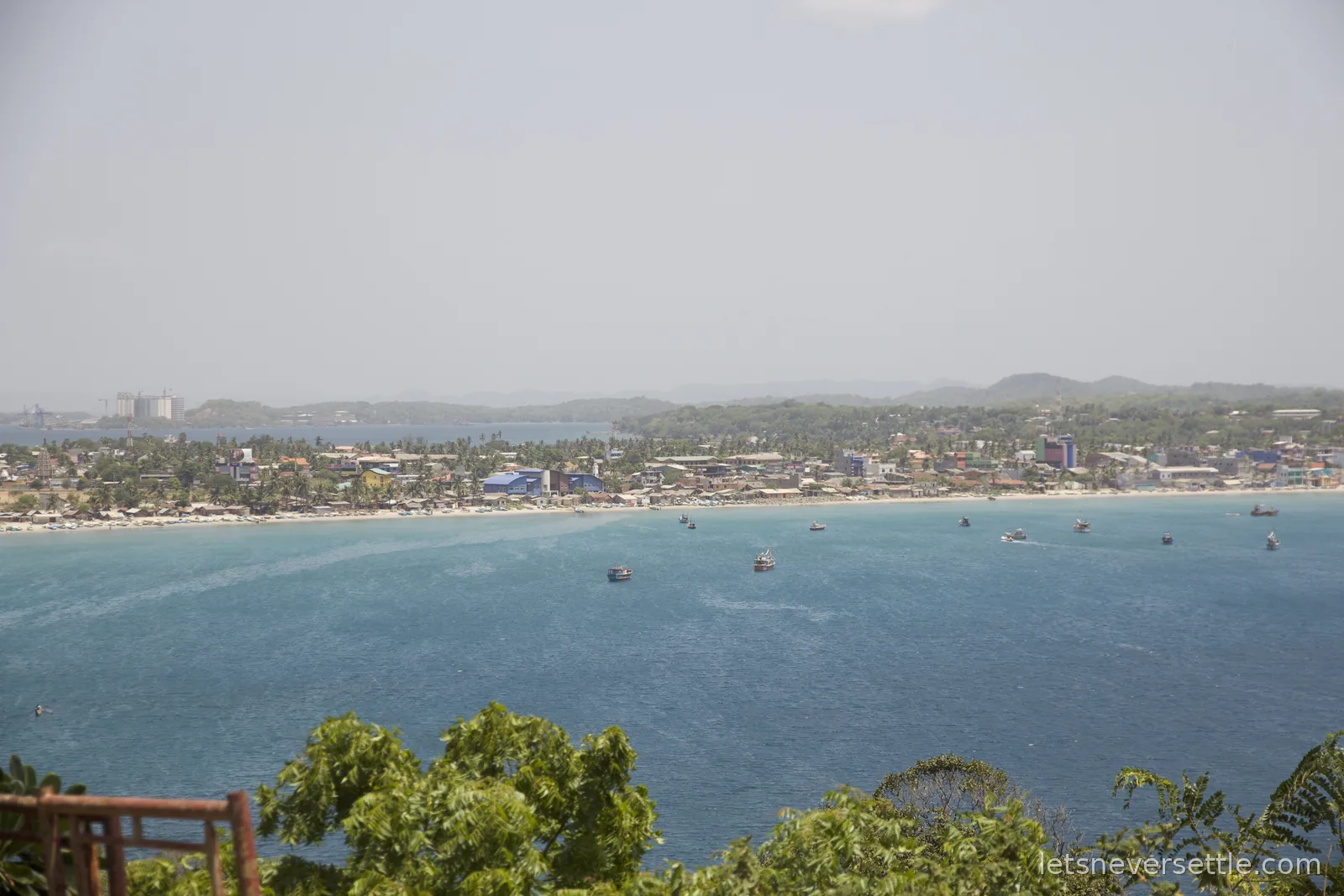 View from Swami Rock over a wide blue bay, Trincomalee town and its beach curving along the far shore with fishing boats on the water