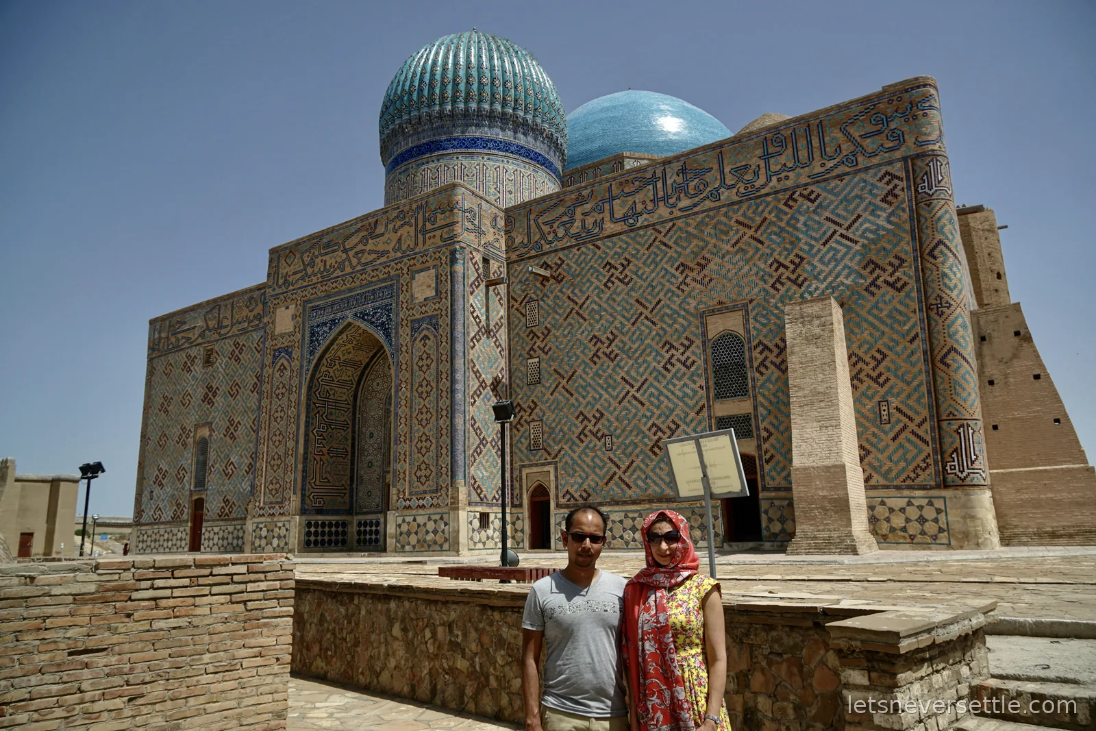 Inga in a red headscarf and Dileep in front of the Khoja Ahmed Yasawi Mausoleum, walls of geometric blue tilework with Kufic script beneath a ribbed turquoise dome