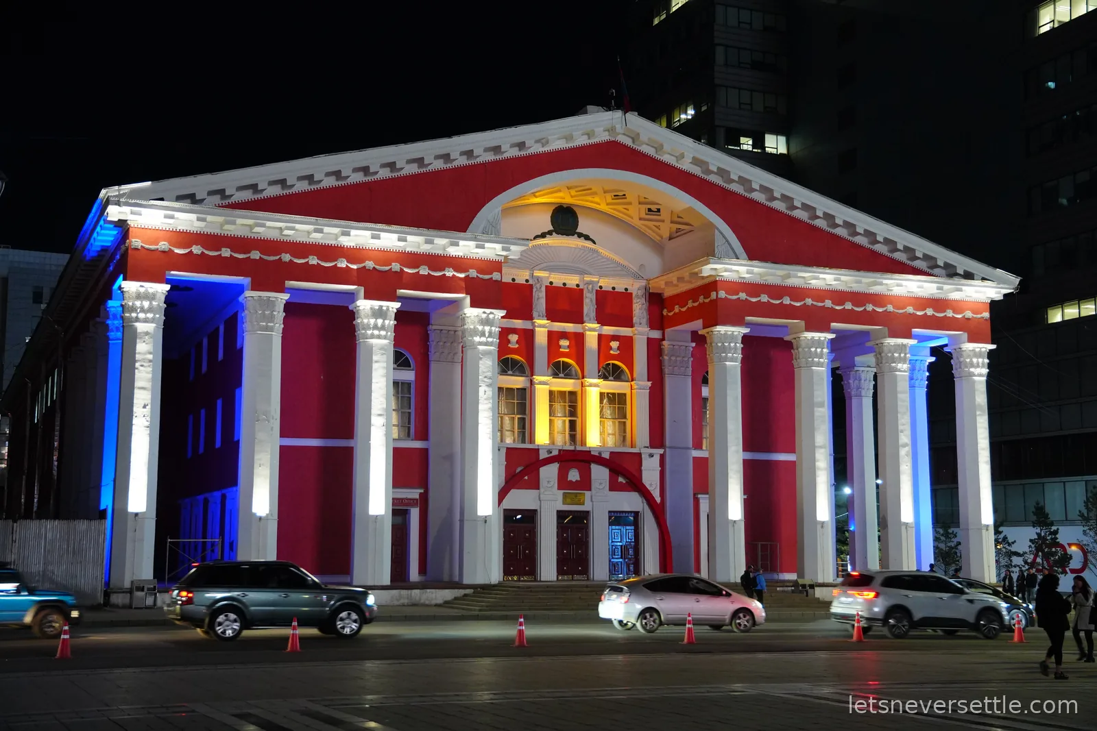 The red neoclassical Opera and Ballet theatre of Ulaanbaatar lit up at night, white columns glowing against the dark office towers behind