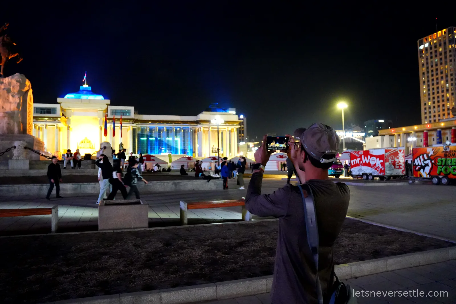 Dileep photographing the illuminated Government Palace across Sukhbaatar Square at night, with street food trucks parked along the square