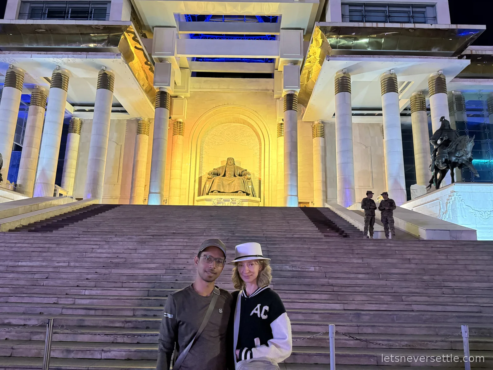 Inga and Dileep at night on the steps of the Government Palace on Sukhbaatar Square, the seated Genghis Khan statue glowing gold behind