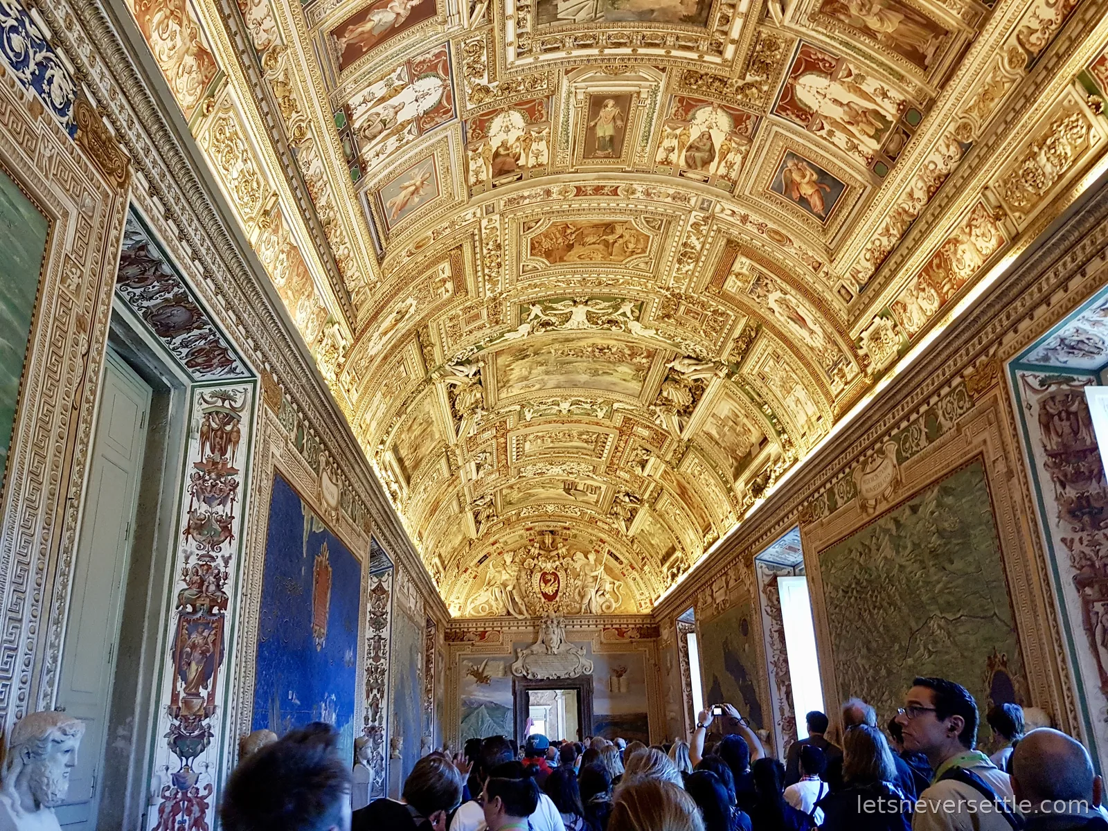 A gallery with a barrel vault covered in gold and painted panels, tapestries on the walls and a dense crowd of visitors below