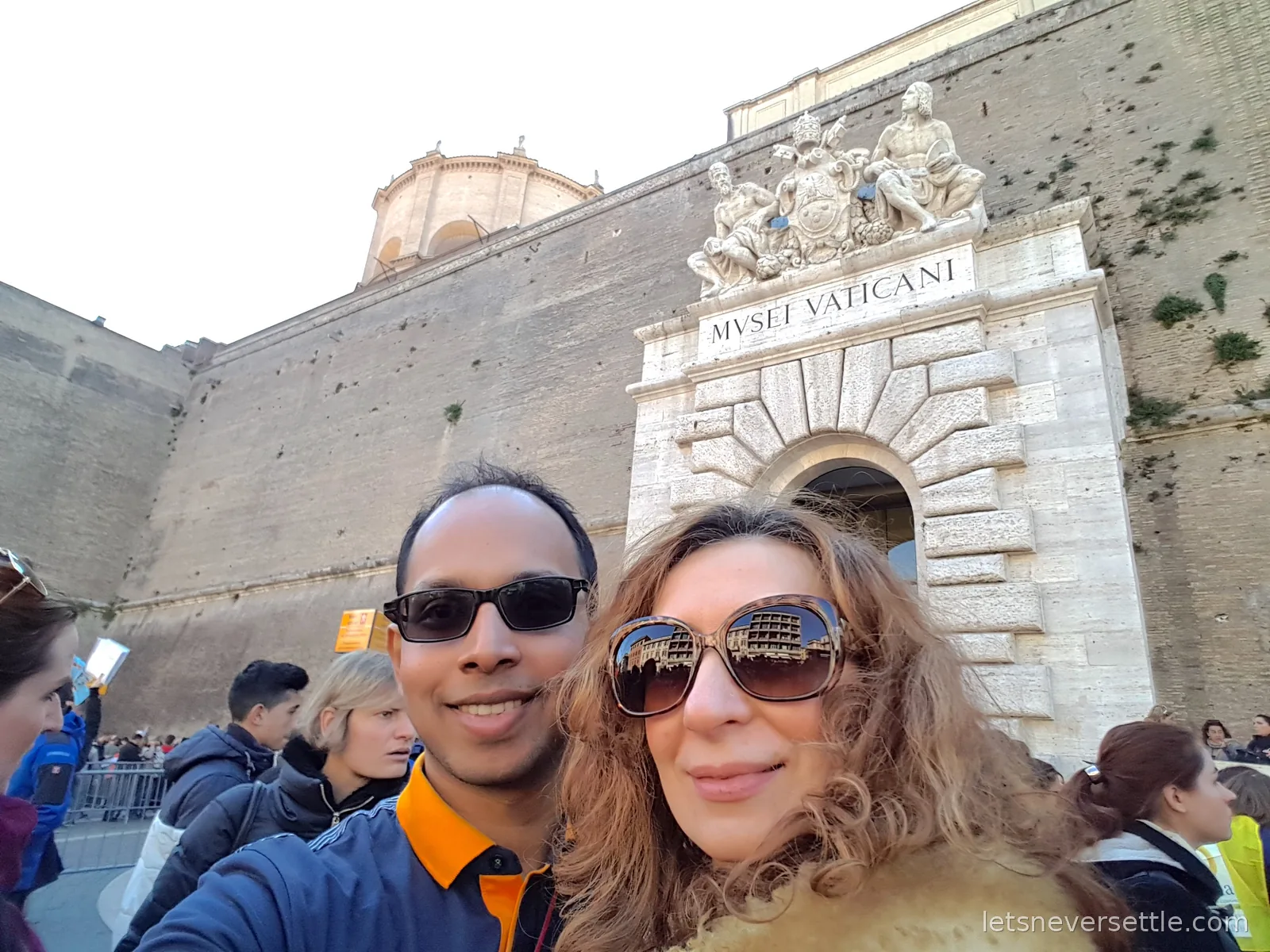 Inga and Dileep taking a selfie in sunglasses below the carved stone doorway of the Vatican Museums, marked Musei Vaticani, with the crowd barriers behind them