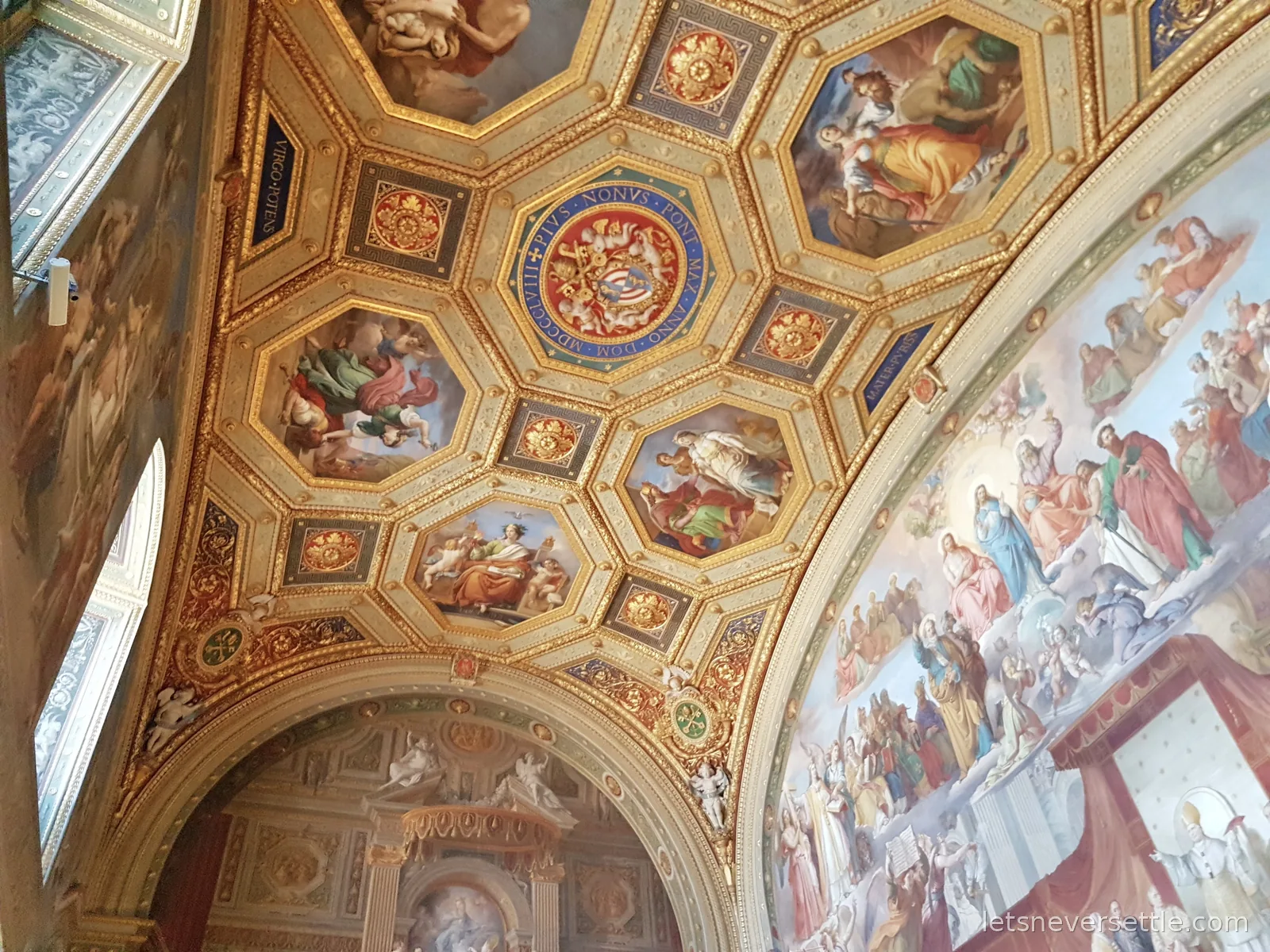 An octagonal coffered ceiling painted with figures and a coat of arms, next to a wall fresco crowded with saints