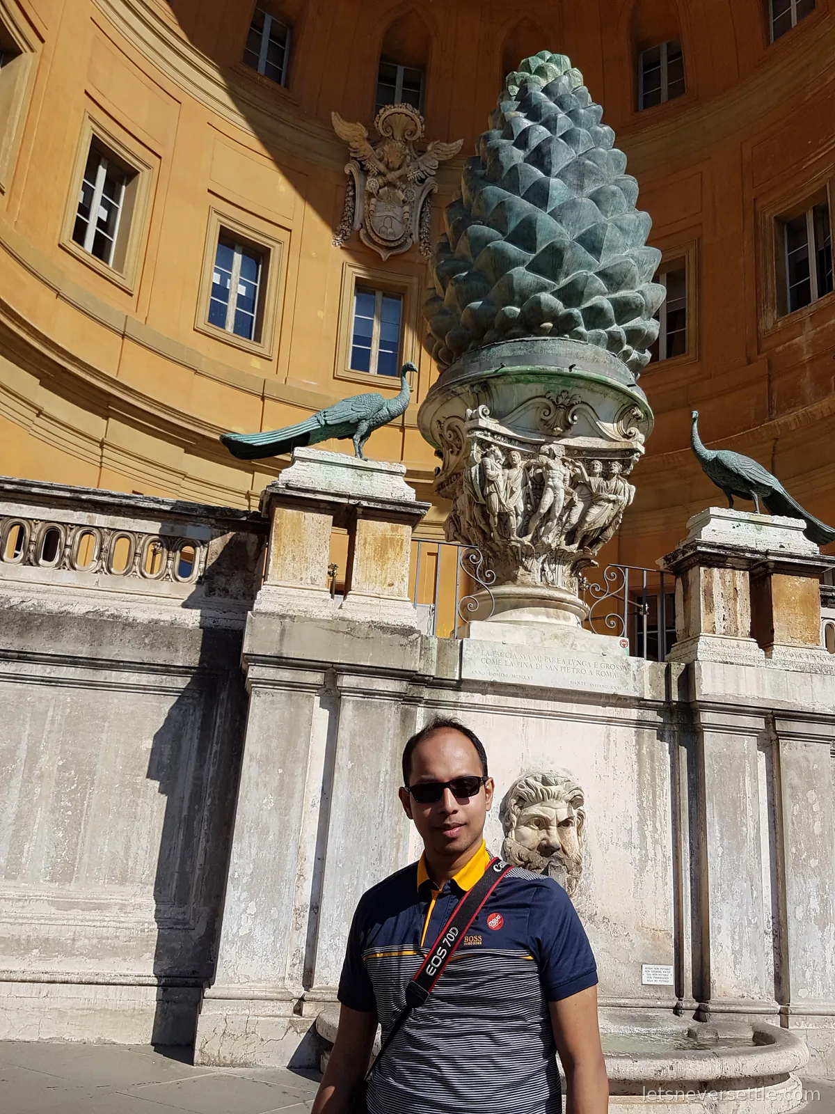 Dileep in sunglasses standing in front of a giant bronze pine cone fountain flanked by two bronze peacocks