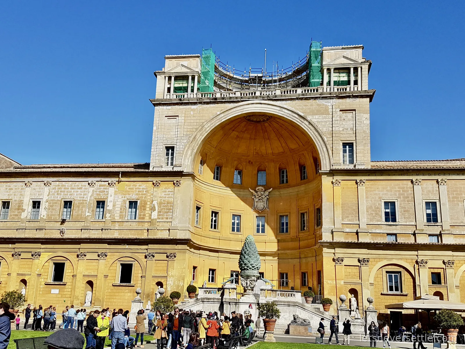 The huge curved niche of the Vatican courtyard in yellow stone, with the bronze pine cone standing at the foot of the steps and crowds in front