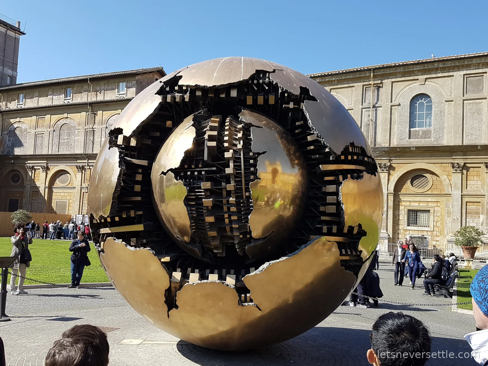 A large bronze sphere sculpture with a broken outer shell revealing a second cracked sphere of gears inside, standing in a courtyard