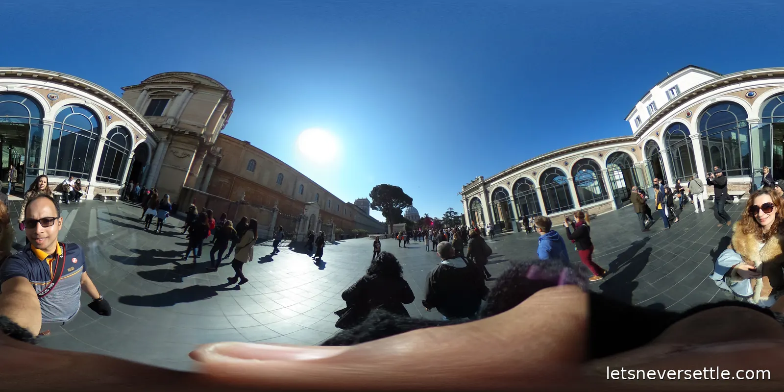 A wide paved museum courtyard in bright sun, edged with tall glazed gallery windows and full of visitors
