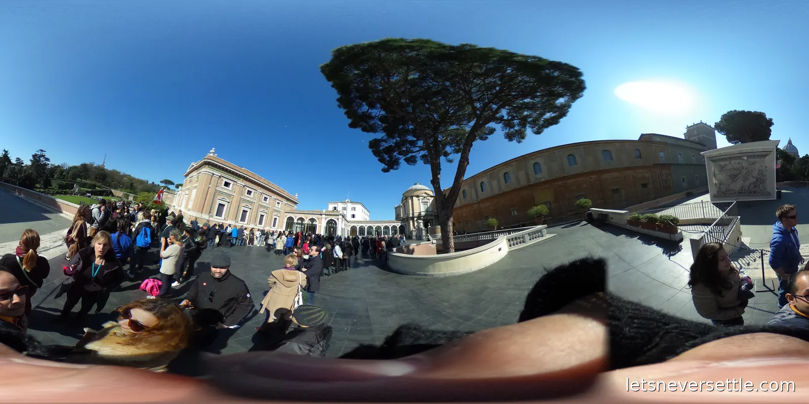 A museum terrace under a large umbrella pine, with a long line of visitors waiting along the wall and the dome of St Peter's beyond