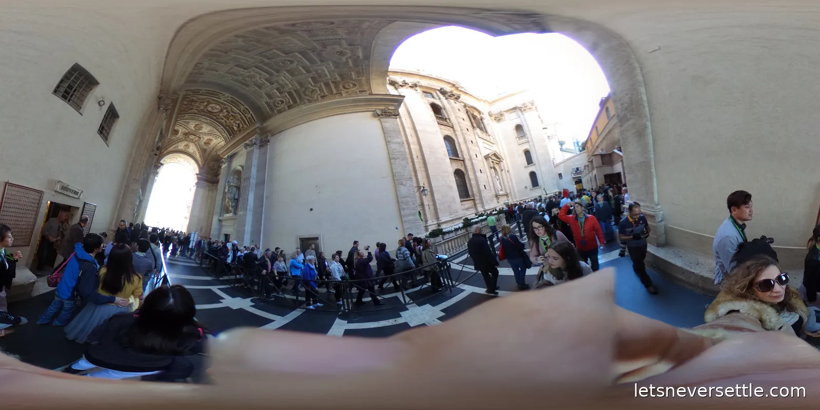 A vaulted stone portico with a patterned floor, opening onto daylight and the side wall of St Peter's Basilica, with visitors queueing