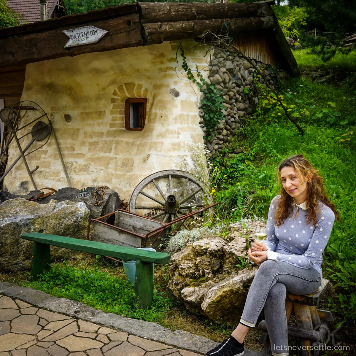 Inga sitting on a low wooden stool with a glass of white wine beside an old whitewashed cottage with a wooden cart wheel, a green bench and a sign reading Kulturny dom