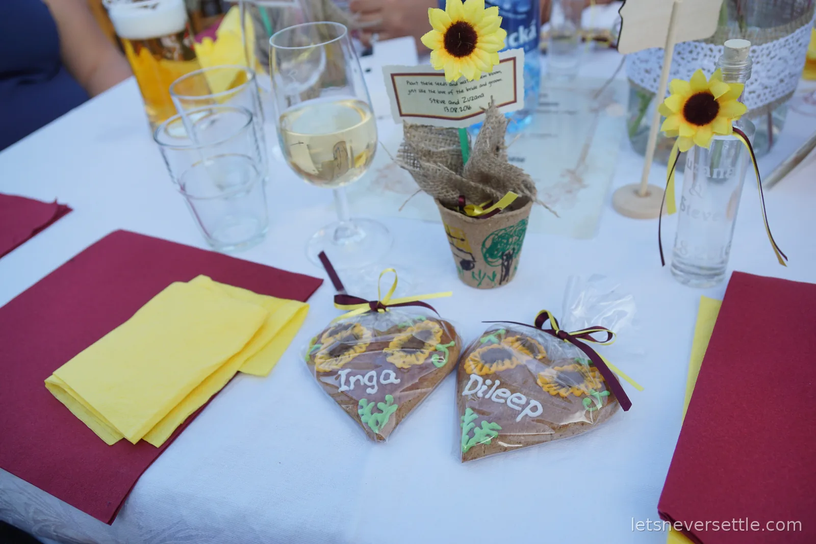 A wedding table set with white cloth, burgundy and yellow napkins, sunflowers in burlap wrapped pots, wine glasses, and two iced gingerbread hearts named Inga and Dileep