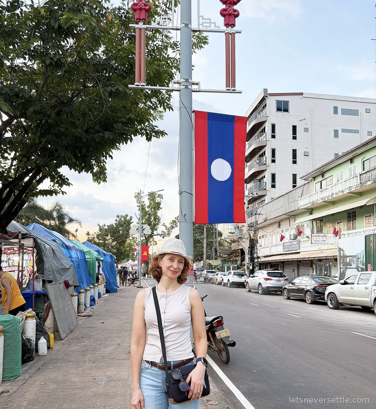 Inga standing on a Vientiane pavement under a hanging Lao flag, with covered night market stalls under blue tarpaulins along the kerb behind her