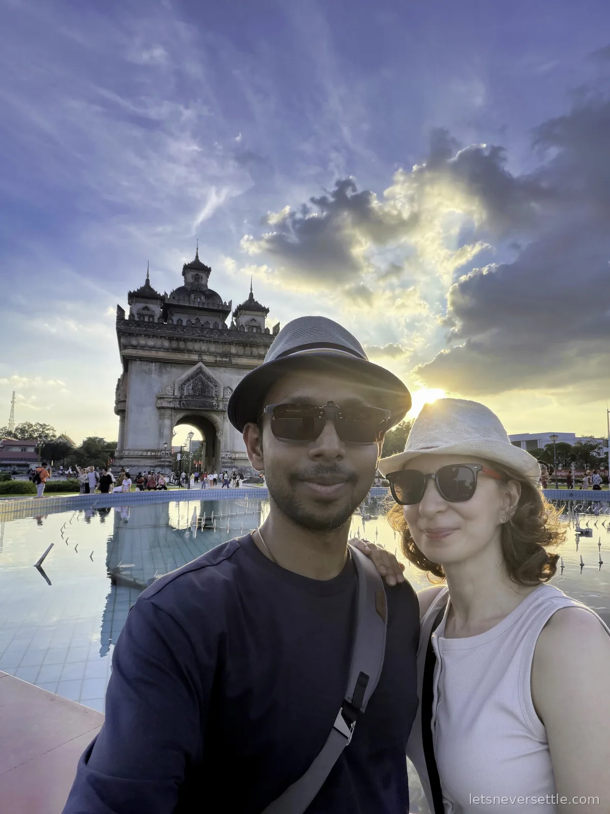 Inga and Dileep in front of the Patuxai victory arch at sunset, the fountain pool reflecting the clouds