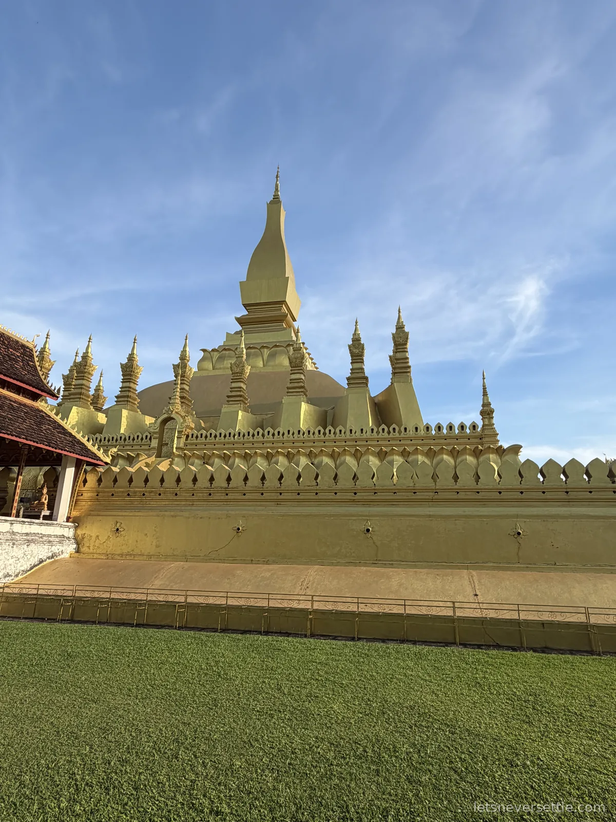 The golden stupa of Pha That Luang rising above its crenellated gold outer wall, seen across mown grass under a blue sky