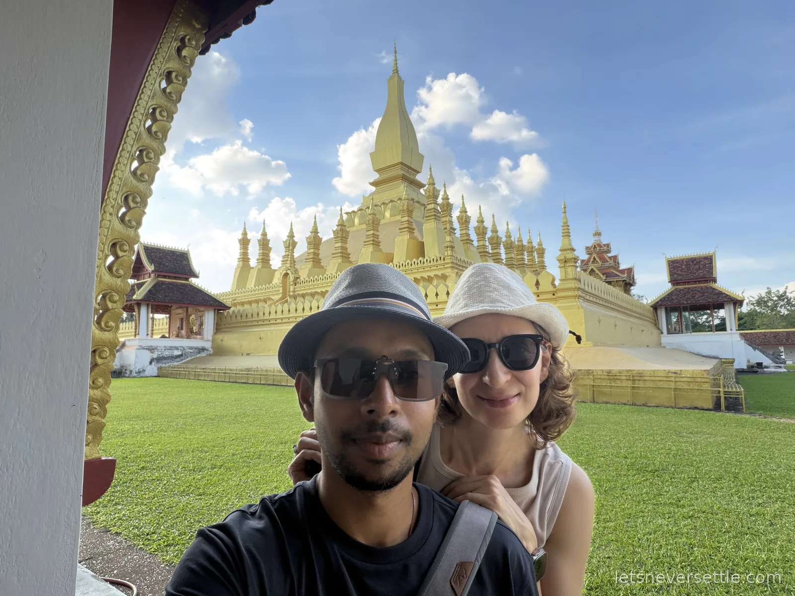 Selfie of Dileep and Inga in sun hats in front of the golden stupa of Pha That Luang under a blue sky