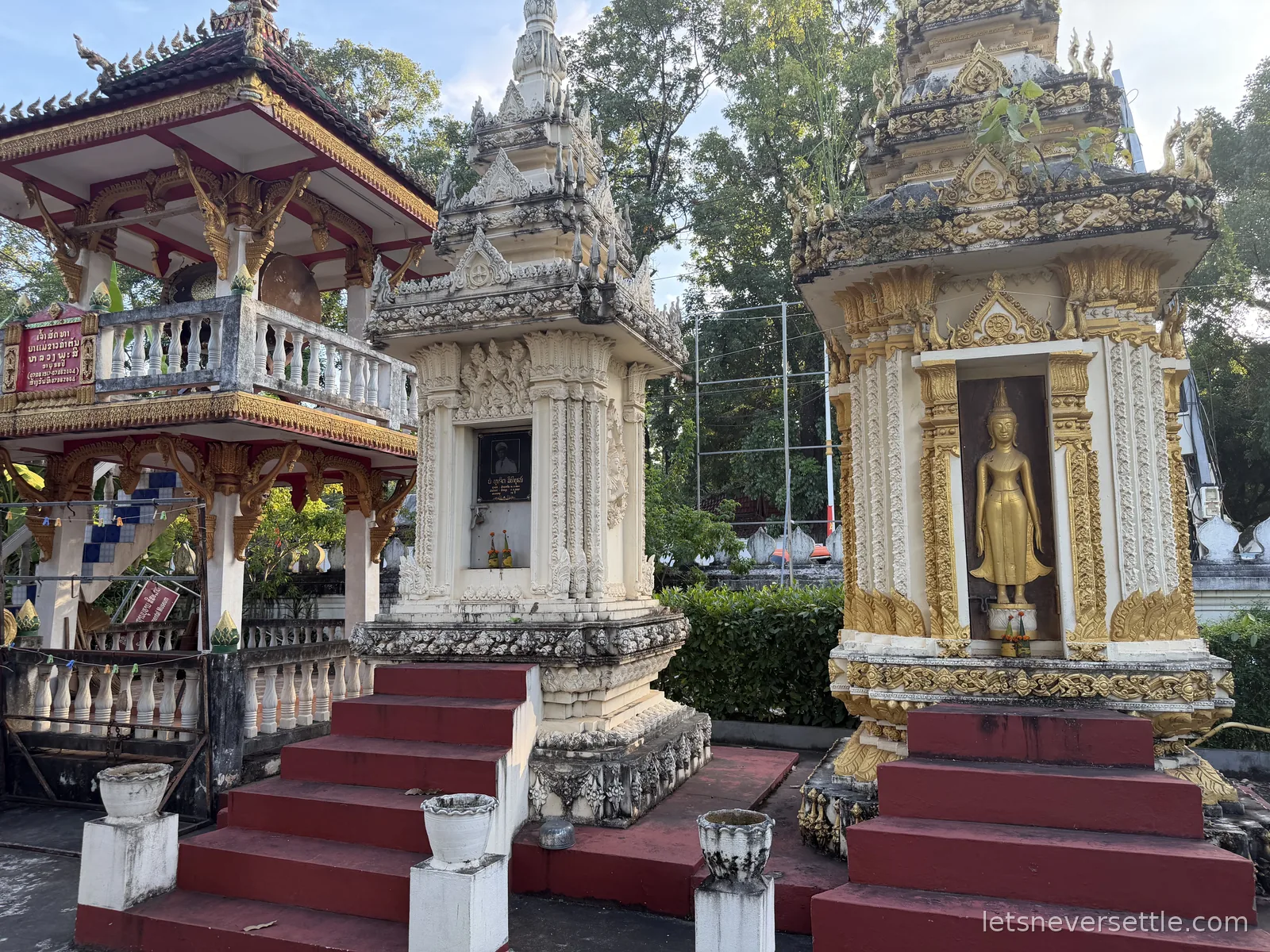 Three ornate white and gold memorial shrines with red-tiled steps beside a temple pavilion, one holding a standing golden Buddha