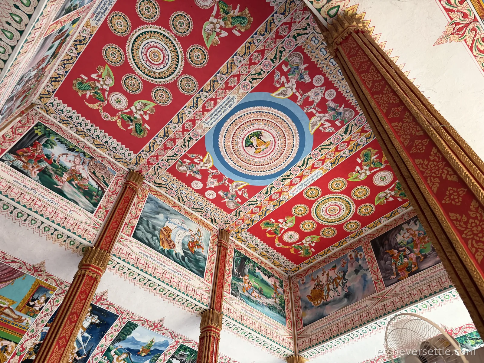 Painted temple ceiling in red and white with gold mandala roundels and rows of framed murals showing scenes from Buddhist stories