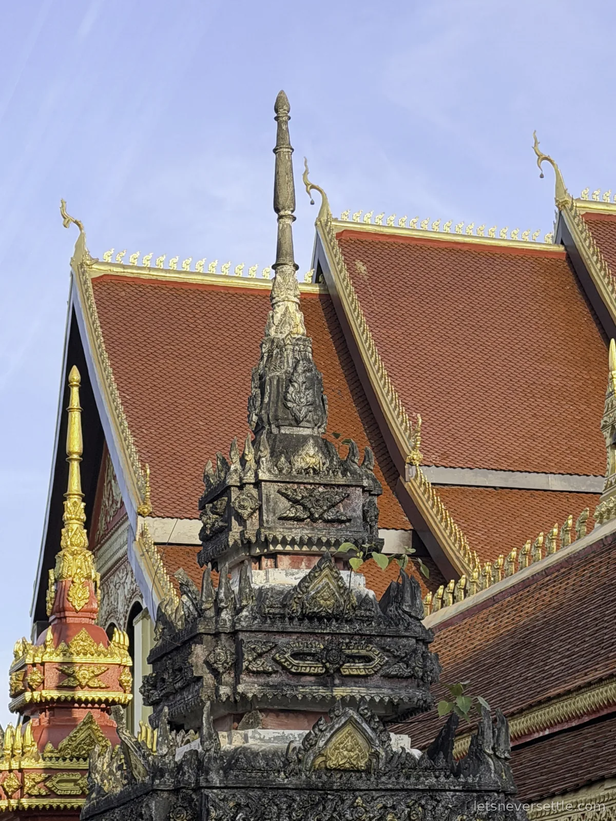 A dark carved stone finial with gold detailing in front of the orange tiled roofs and gilded eaves of a Lao temple