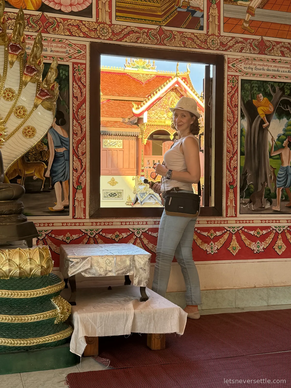 Inga in a white sun hat standing beside an open temple window framed by painted murals, another gilded temple roof visible through the opening