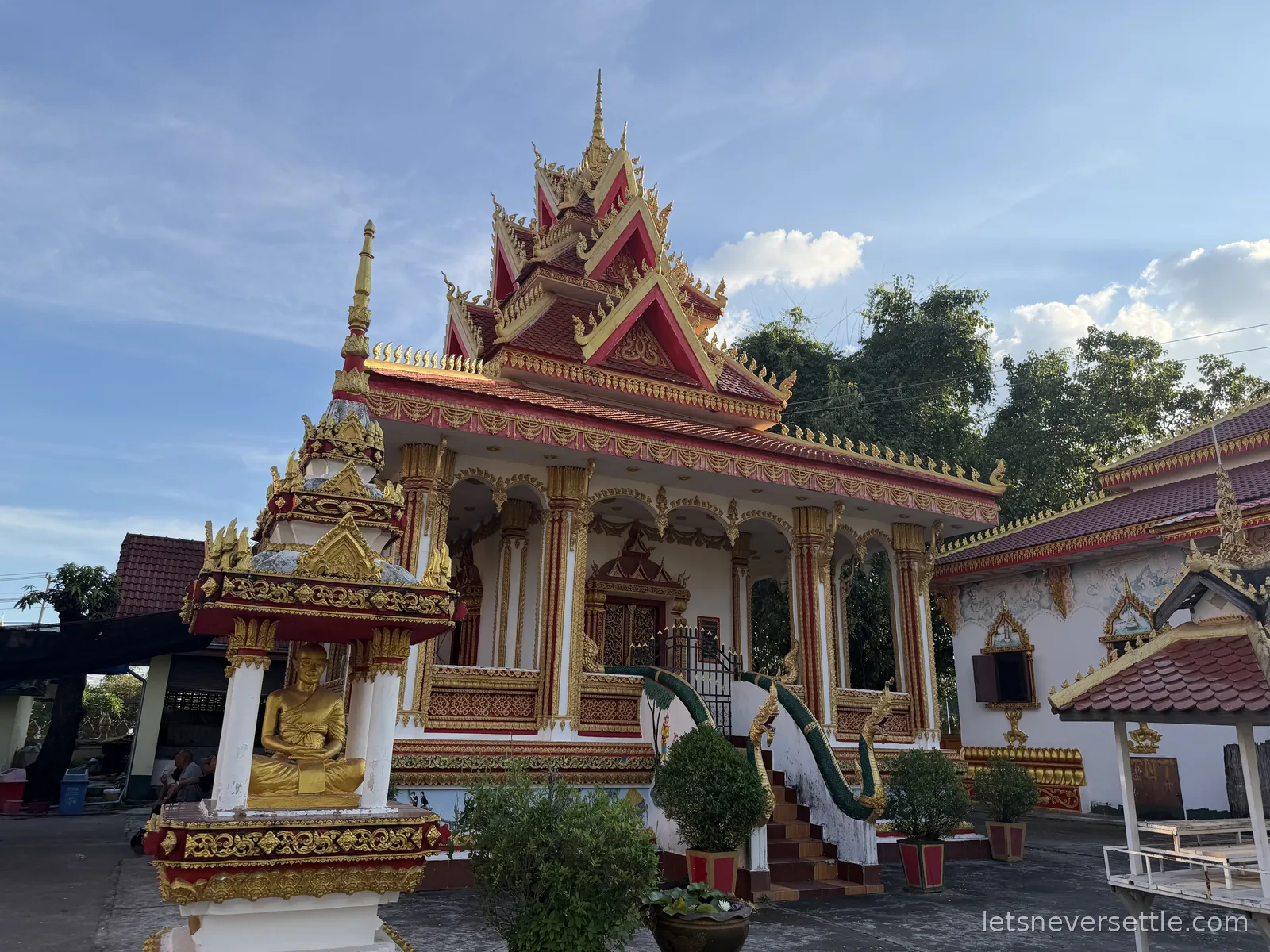 A Lao temple hall with a red and gold tiered roof, ornate gilded columns and a golden Buddha in a small roadside shrine in front
