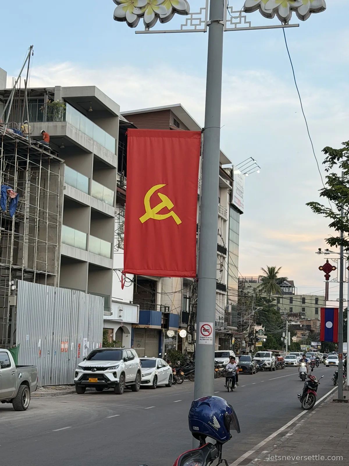 A Vientiane street with a red hammer and sickle banner and the Lao national flag hanging from lamp posts, scaffolding on a half-built block and motorbikes in the traffic