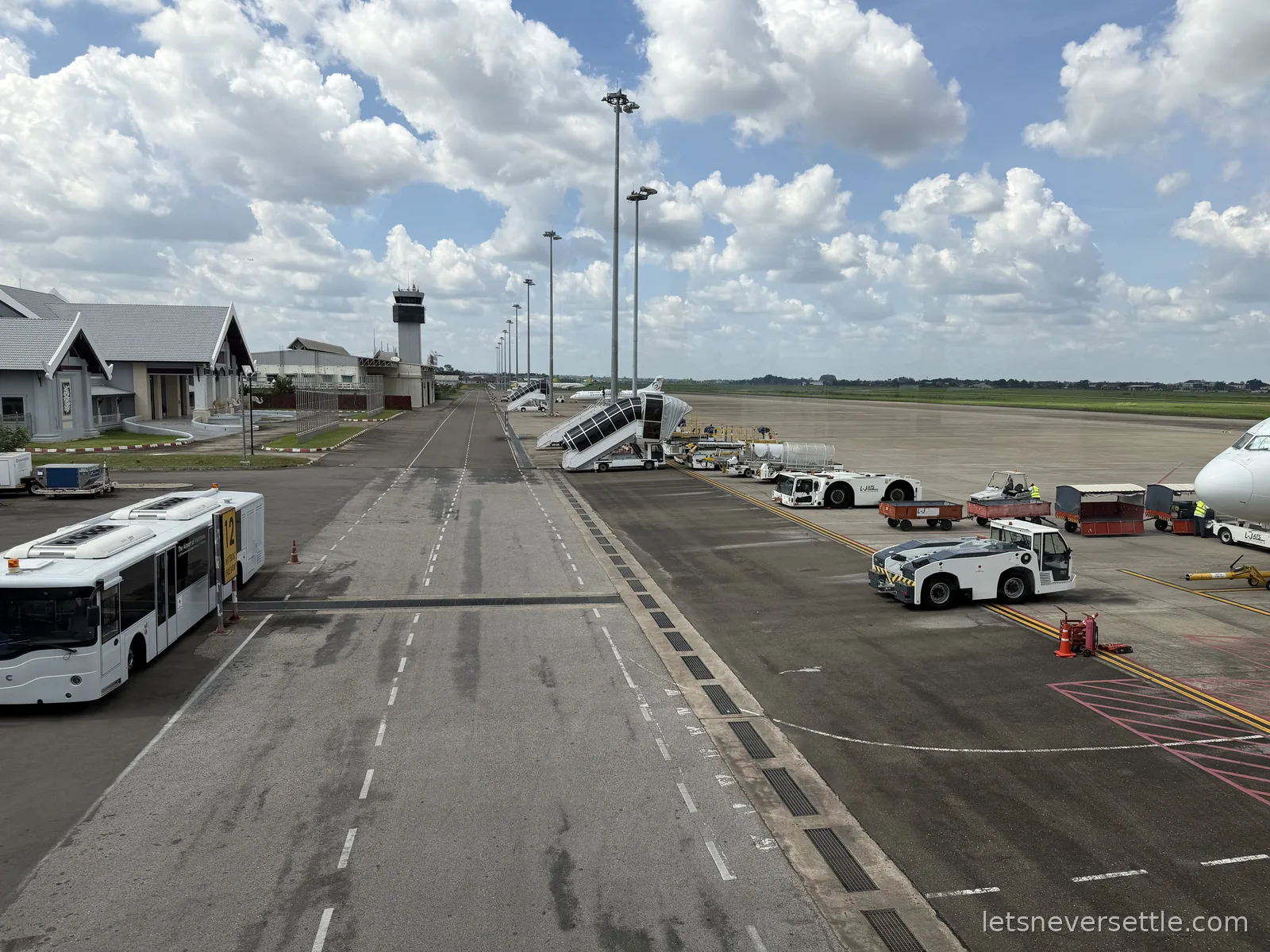 View from a terminal window over the apron at Wattay airport in Vientiane, with airstairs, ground vehicles, a shuttle bus and the control tower under piled white clouds