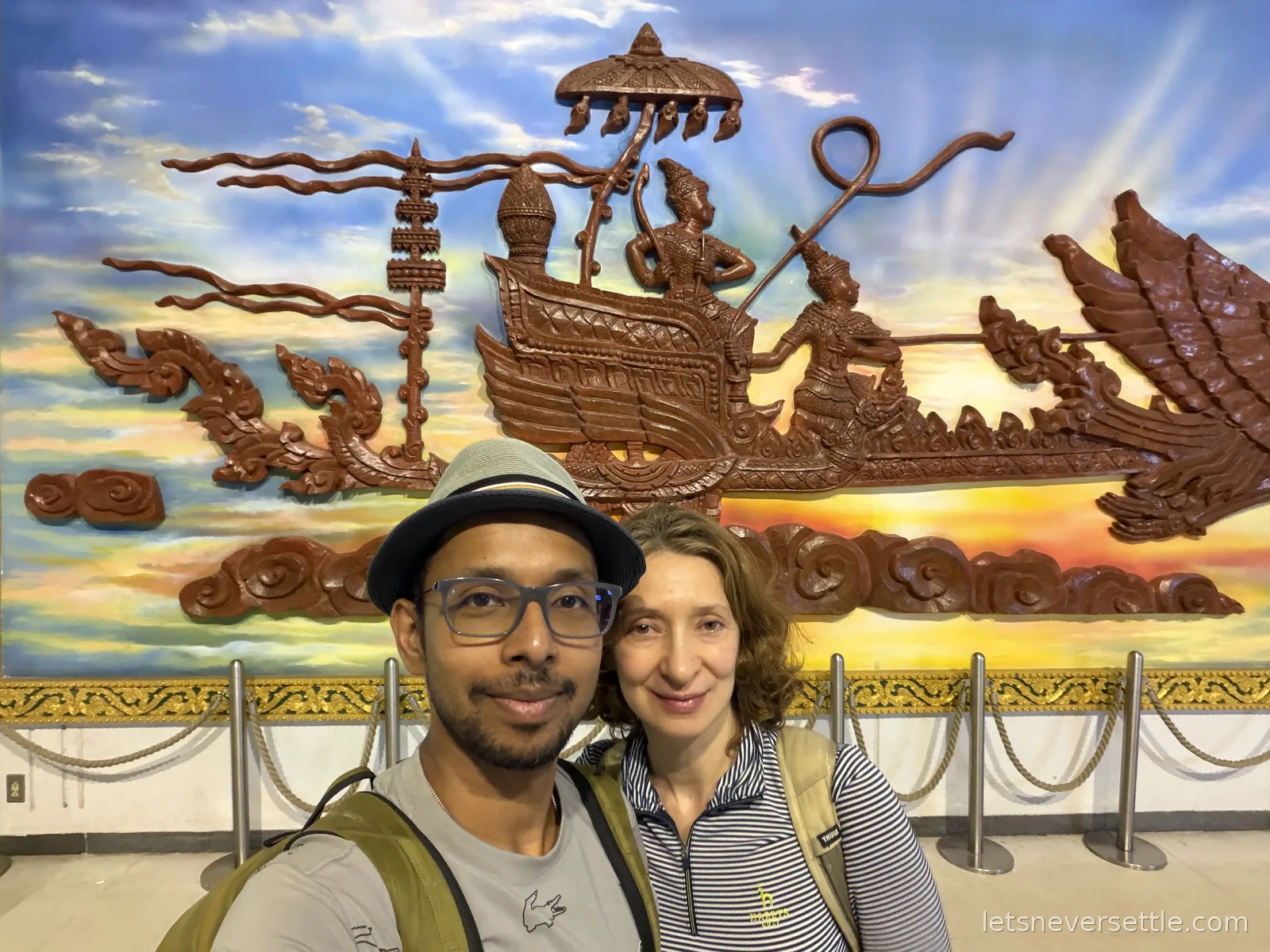 Selfie of Dileep and Inga in front of a large carved wooden relief of a royal barge with mythical serpents, on a painted sunset background at Wattay airport