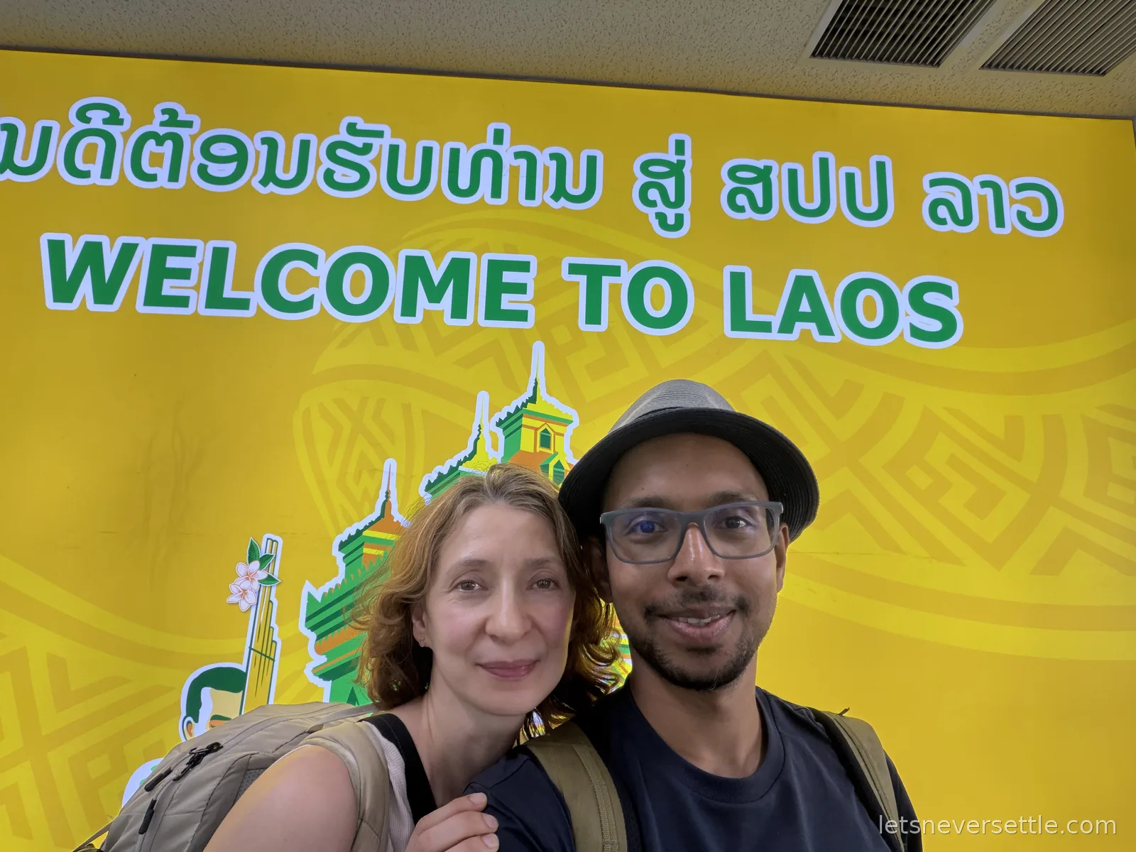 Inga and Dileep taking a selfie in front of a bright yellow Welcome to Laos sign in the arrivals hall