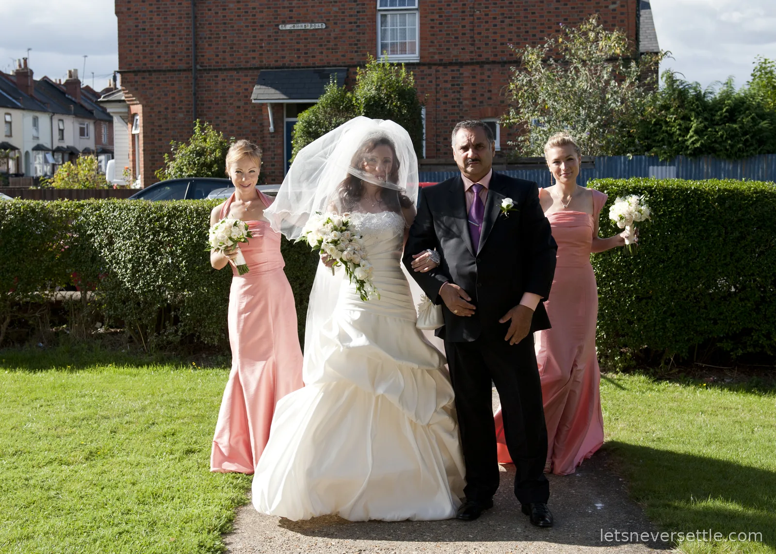 Inga in her veil and wedding dress walking arm in arm along a garden path, with the two bridesmaids in pink either side