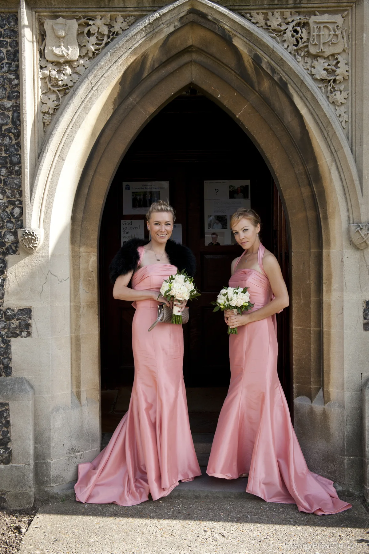 The two bridesmaids in long pink dresses holding white bouquets, framed by the carved stone arch of the church door