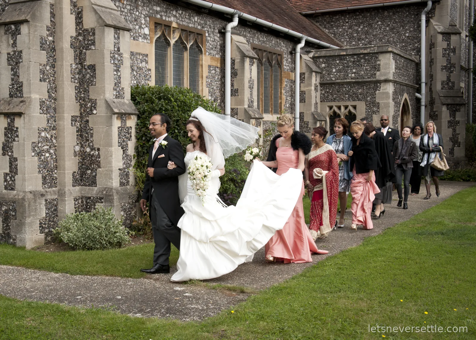 Inga in her veil on Dileep's arm leading the wedding procession along the church path, guests in saris and suits behind