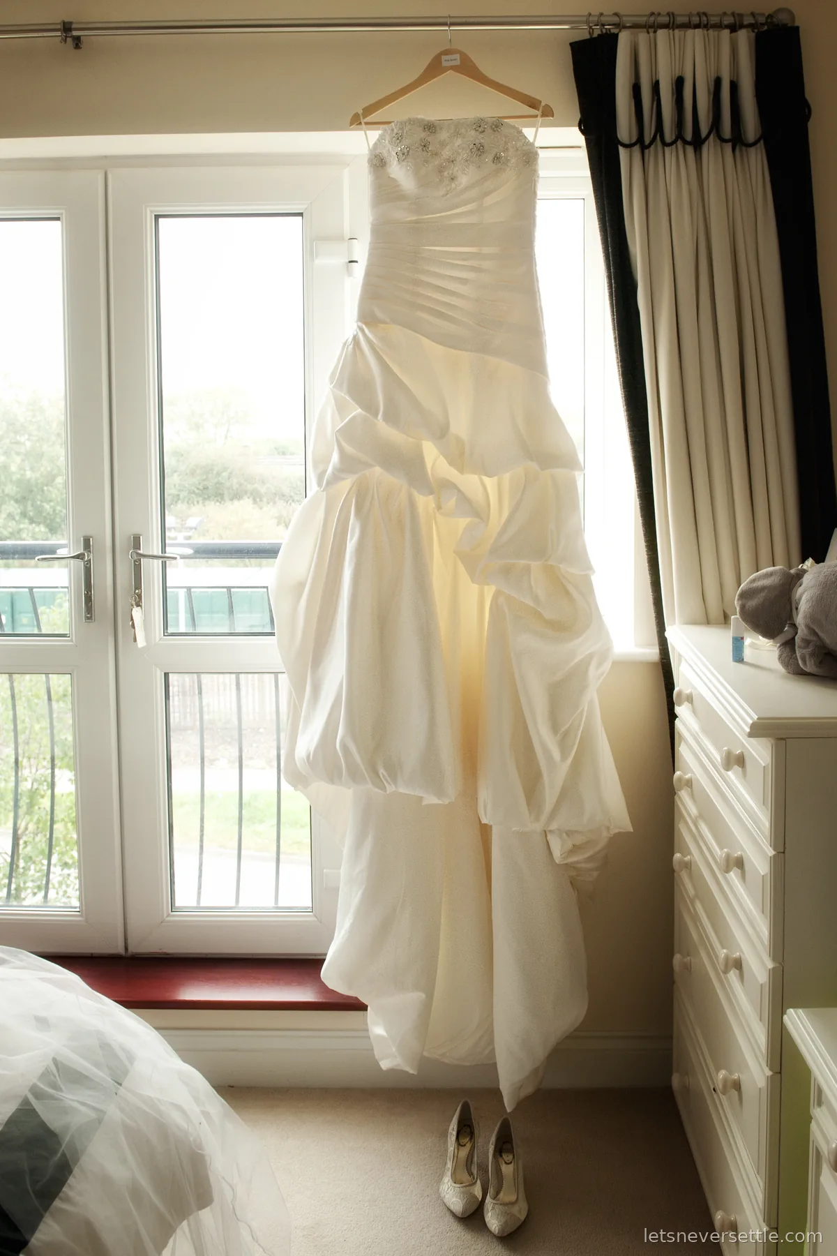 Ivory wedding dress on a wooden hanger against a bright window, with the shoes placed on the carpet below