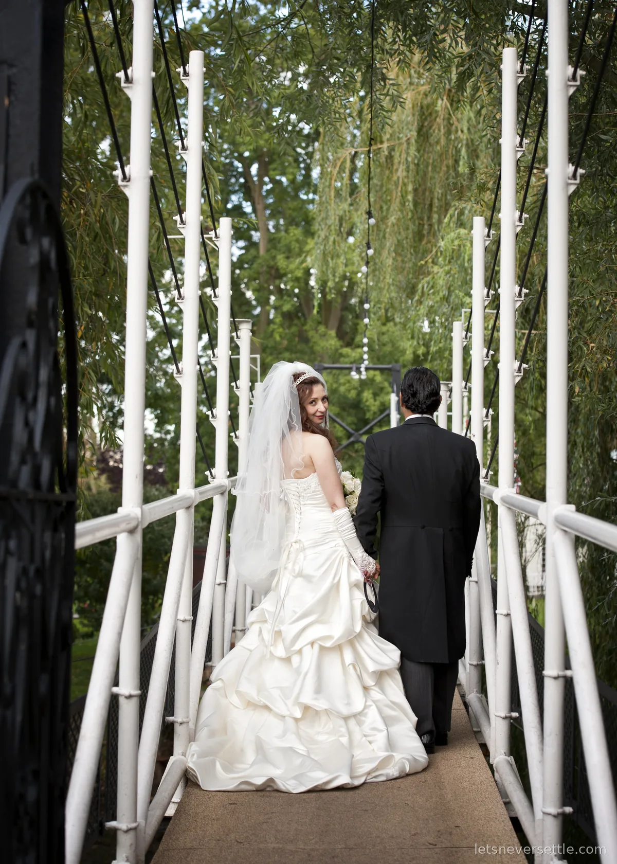 Inga looking back over her shoulder as she and Dileep cross a white suspension footbridge under willow trees