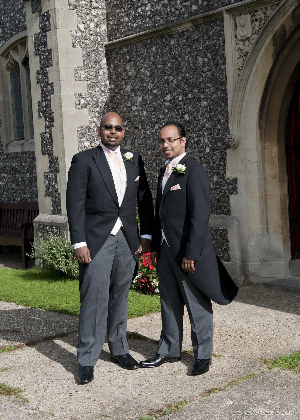 Dileep and his brother in black morning tailcoats with pale pink ties and white rose buttonholes, standing by the flint church wall