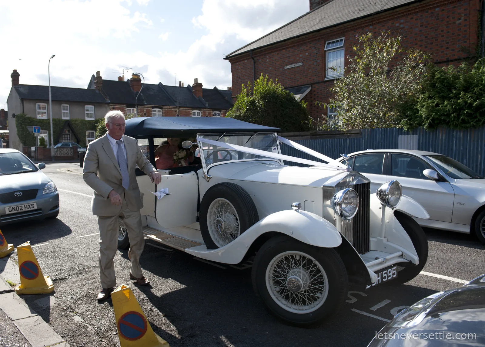 A white vintage Rolls-Royce dressed with white wedding ribbons pulling up on a street of red brick terraced houses