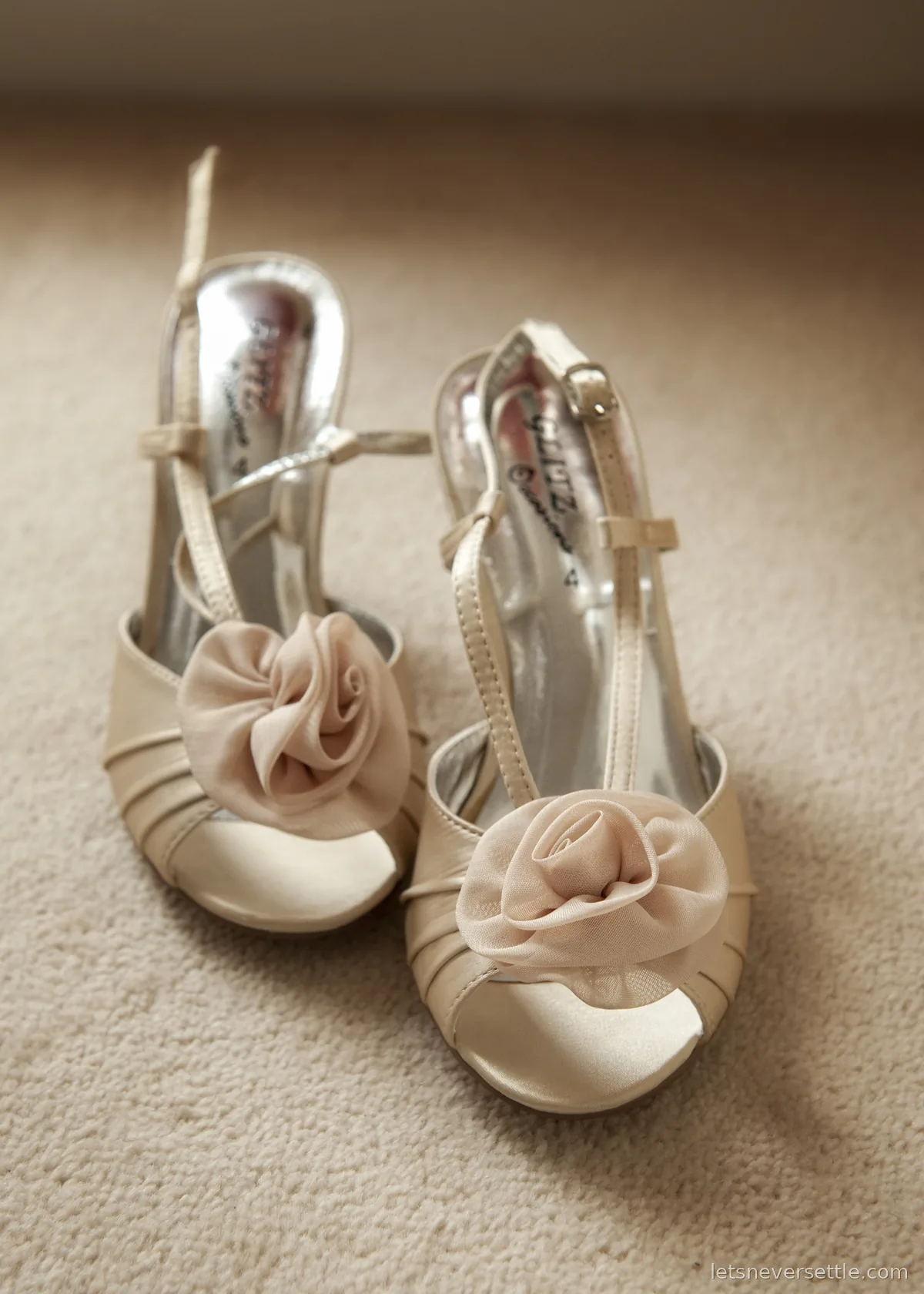 A pair of ivory satin peep-toe wedding shoes with chiffon roses on the toes, set on cream carpet