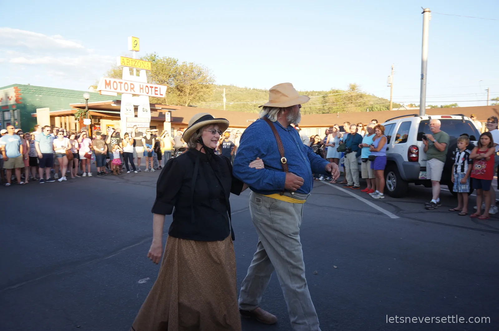 Two actors in Wild West costume walking arm in arm past a crowd gathered outside a motor hotel in Williams