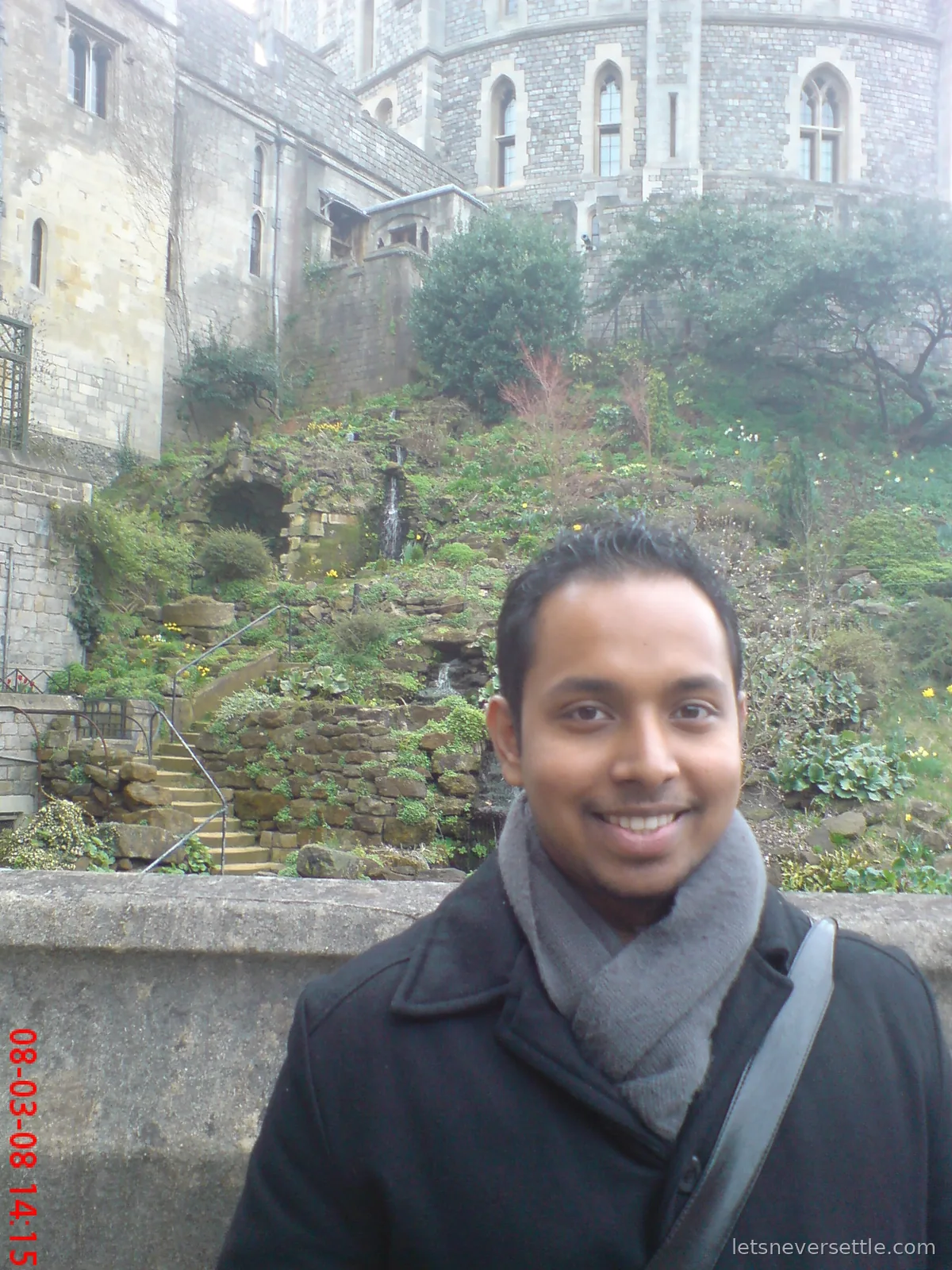 Dileep in a black coat and grey scarf smiling below the Round Tower of Windsor Castle, a rock garden with stone steps and daffodils behind