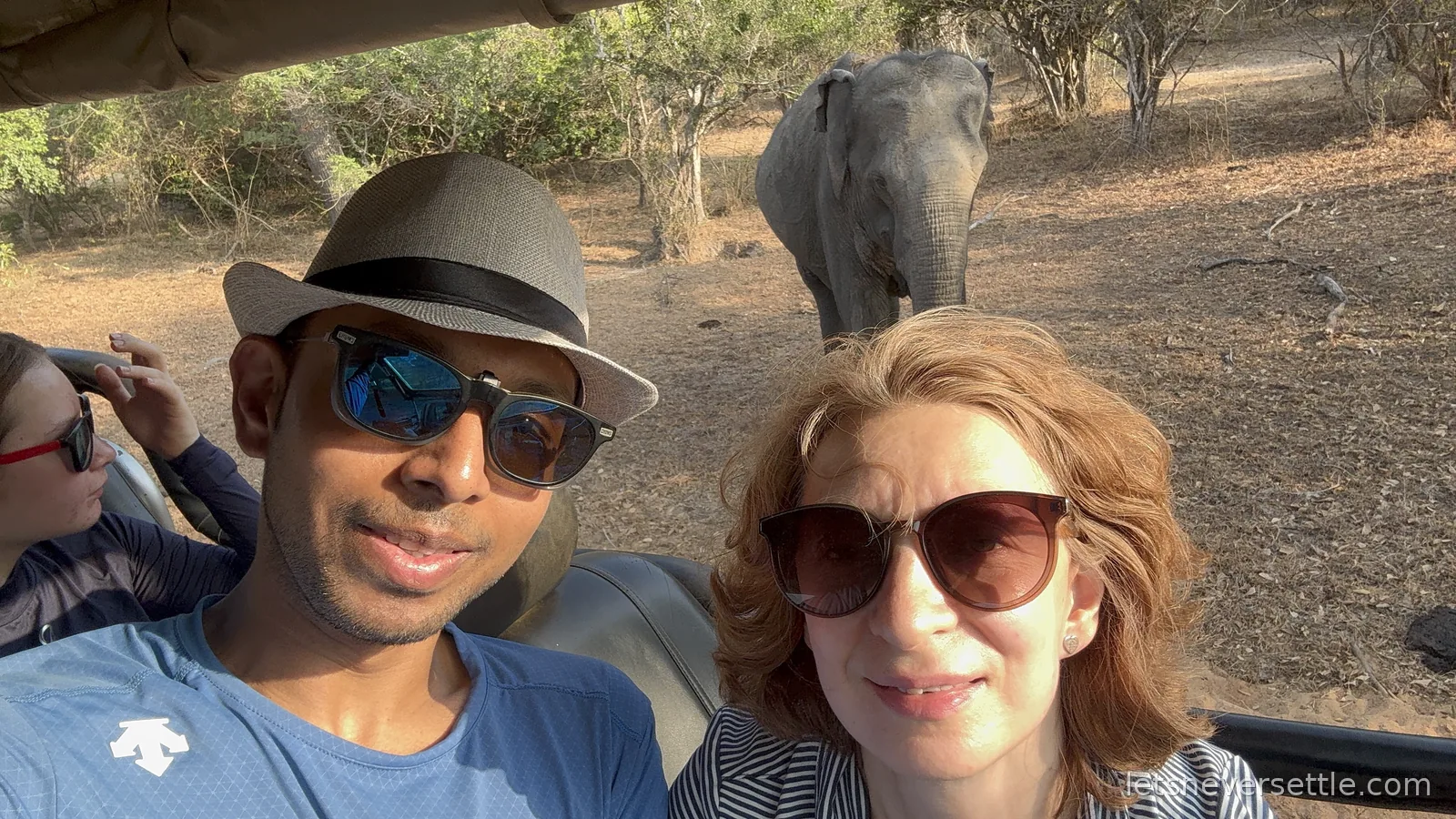 Inga and Dileep taking a selfie in an open safari jeep while a wild elephant walks up the track behind them