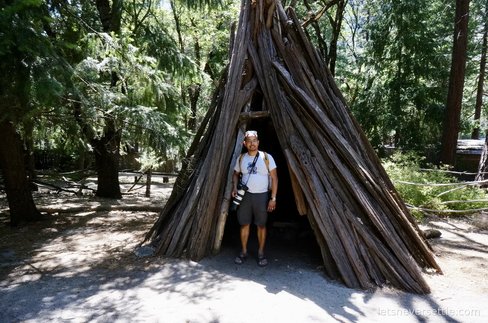Dileep standing in the doorway of a cone shaped cedar bark house among tall trees in Yosemite Valley