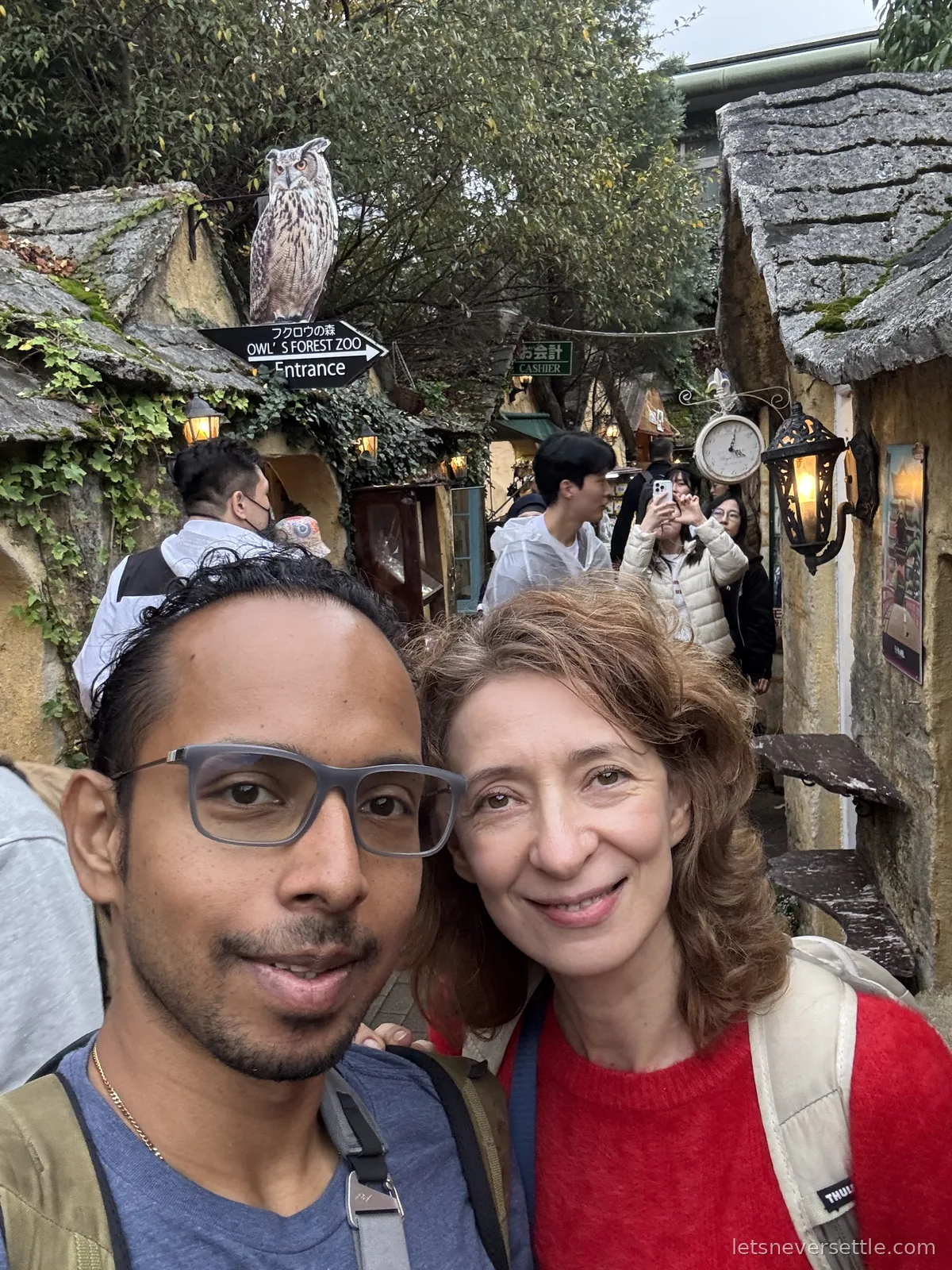 Inga and Dileep taking a selfie in a narrow lane of stone fairy-tale cottages, under an owl figure and a sign for the Owl's Forest Zoo