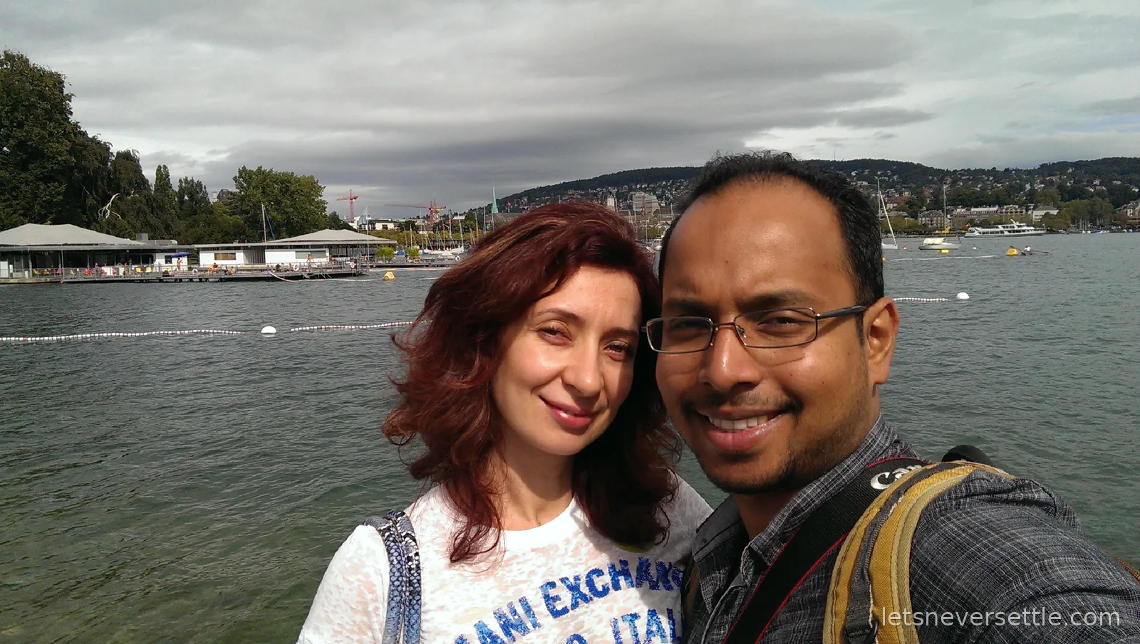 Inga and Dileep selfie on the shore of Lake Zurich with a lakeside lido and hills behind